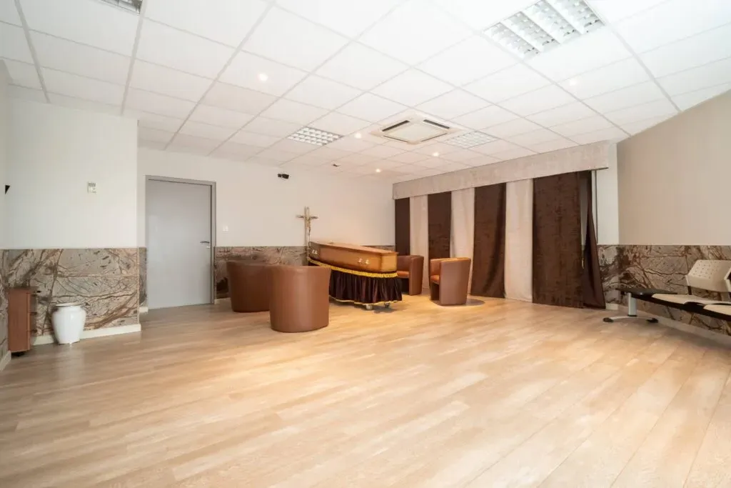 Salon funéraire avec un cercueil sur un catafalque orné et des fauteuils.